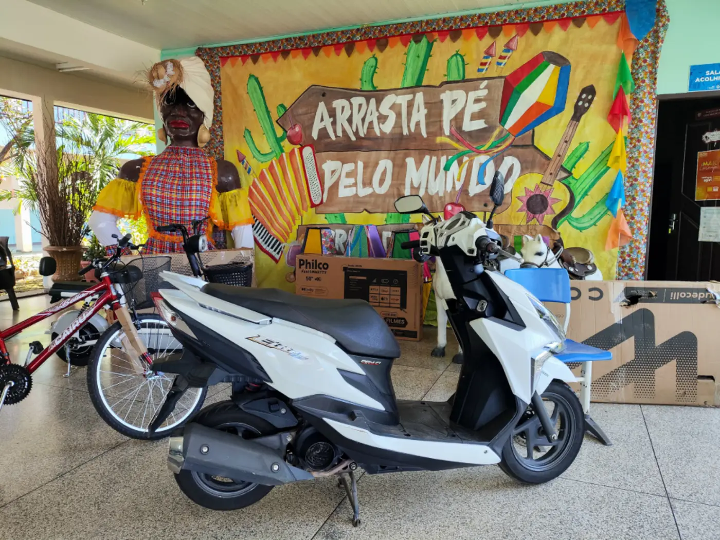 Prêmios do bingo, como moto, TV 4K e bicicletas, estão expostos diante do painel temático ‘Arrasta-pé pelo mundo’, na entrada da Escola Estadual Mário Quirino.