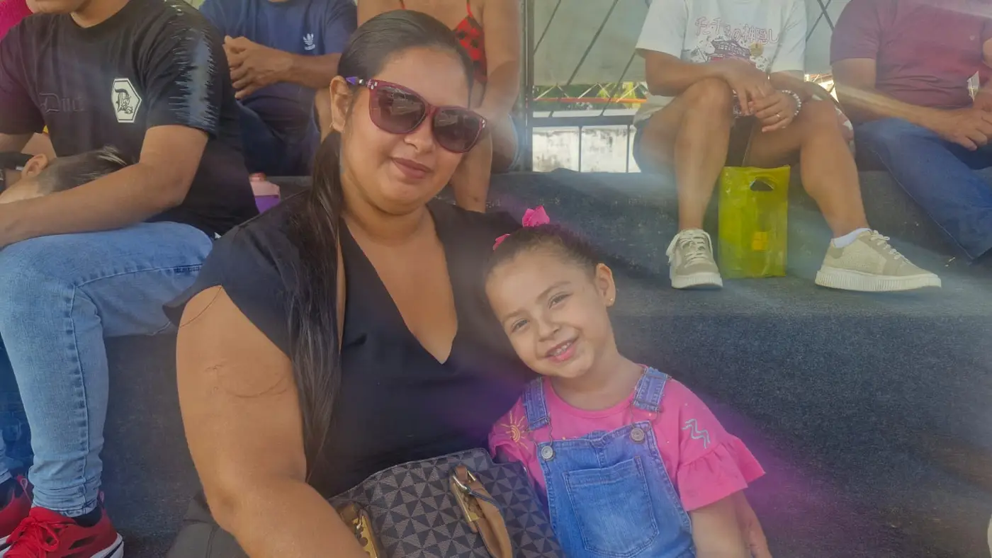 Camila Reis, ao lado da filha Helena, espera com orgulho o desfile do filho Bruno
